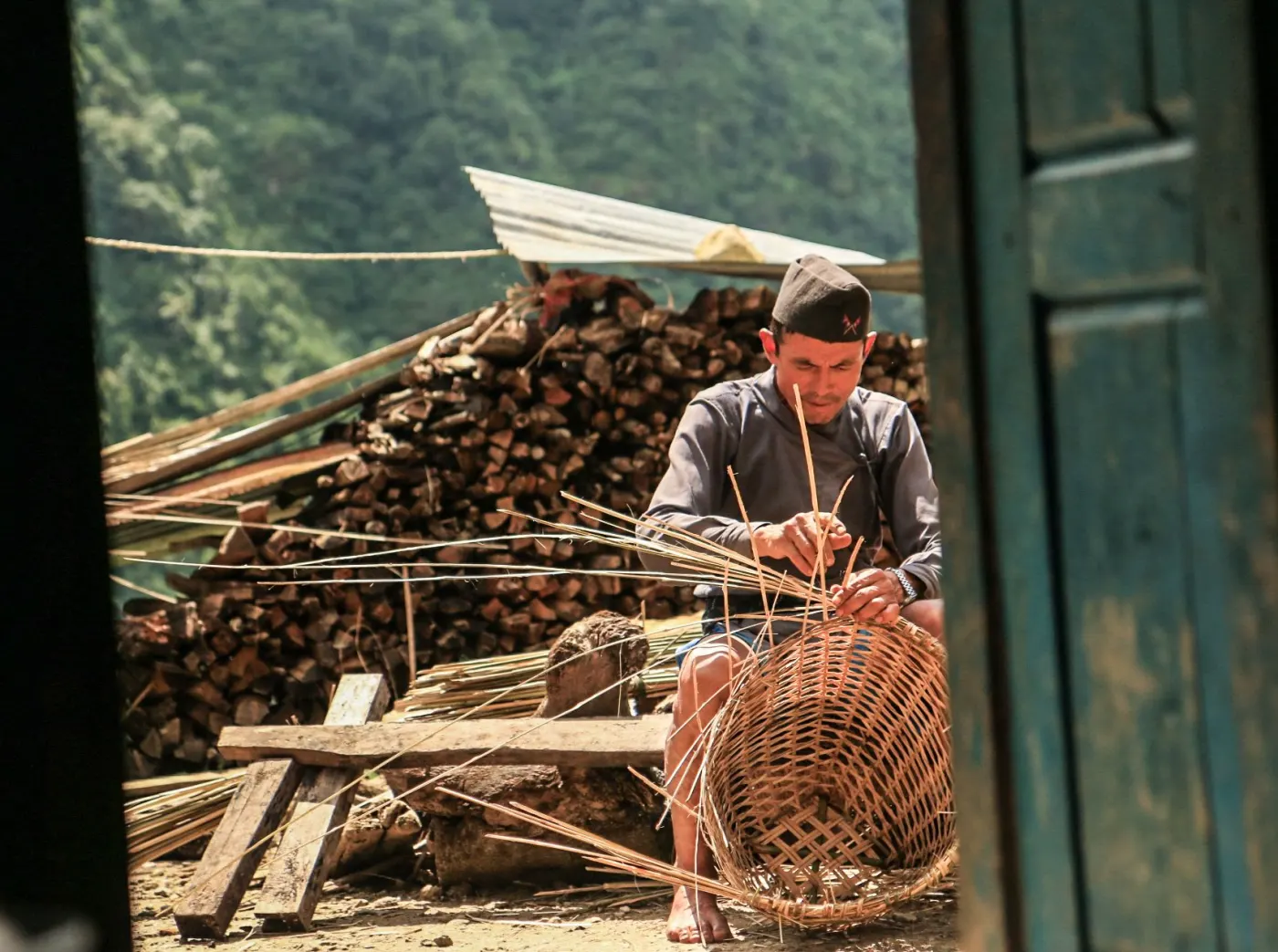 A man weaving a basket beside a stack of firewood in Gyangphedi